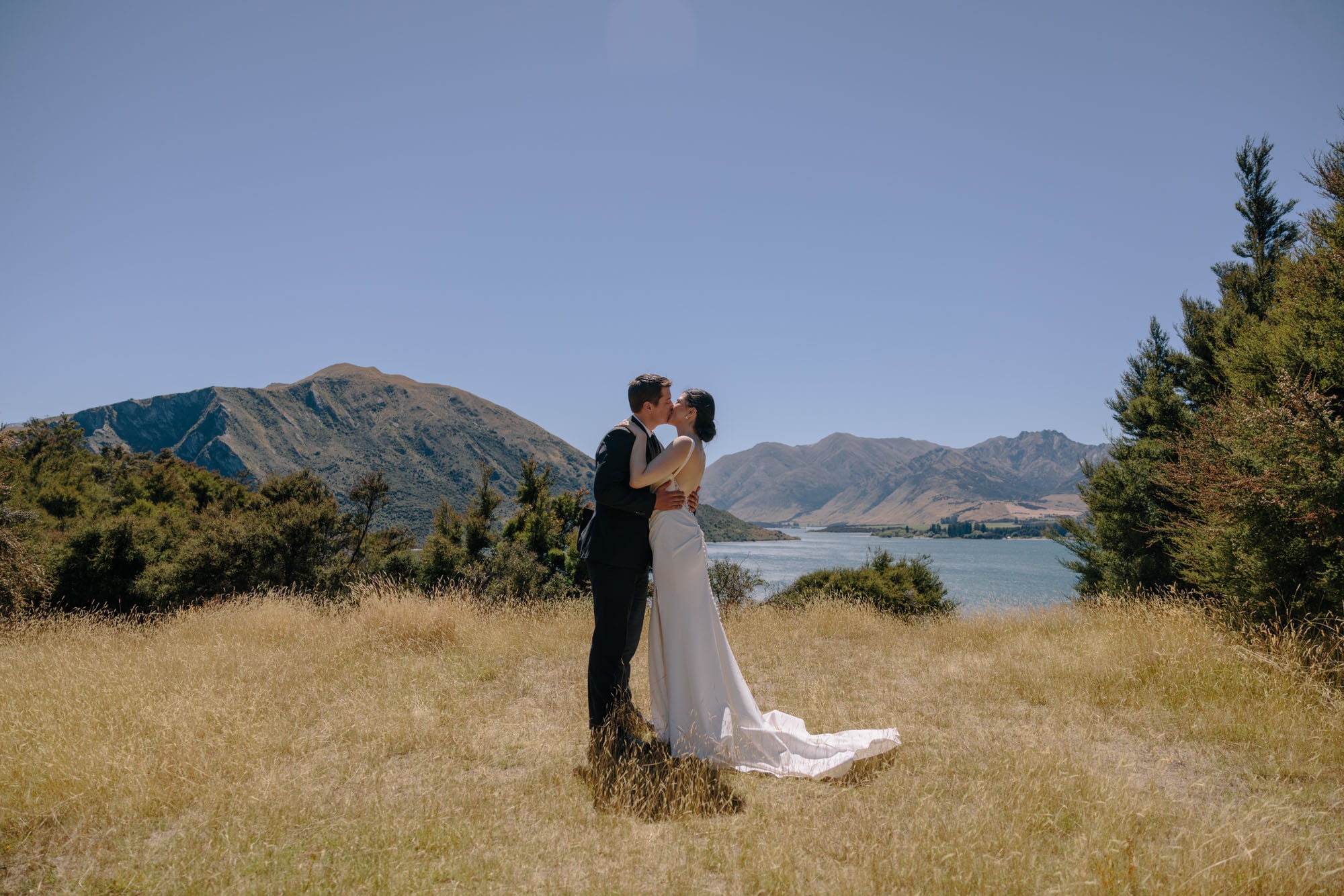 A couple kissing in golden tussock above Lake Wānaka, the mountains across the water behind them