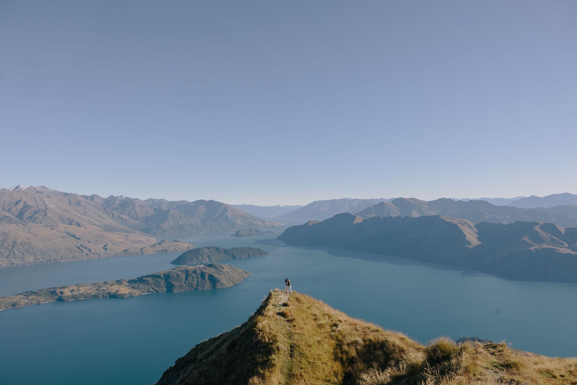 A couple standing at the end of a ridge high above Lake Wānaka, tiny against the water and the mountains beyond